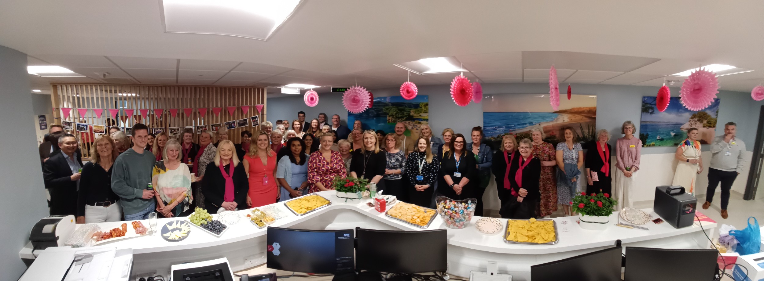 Group of people in hospital unit reception area with buffet food on counter top and bunting on walls.