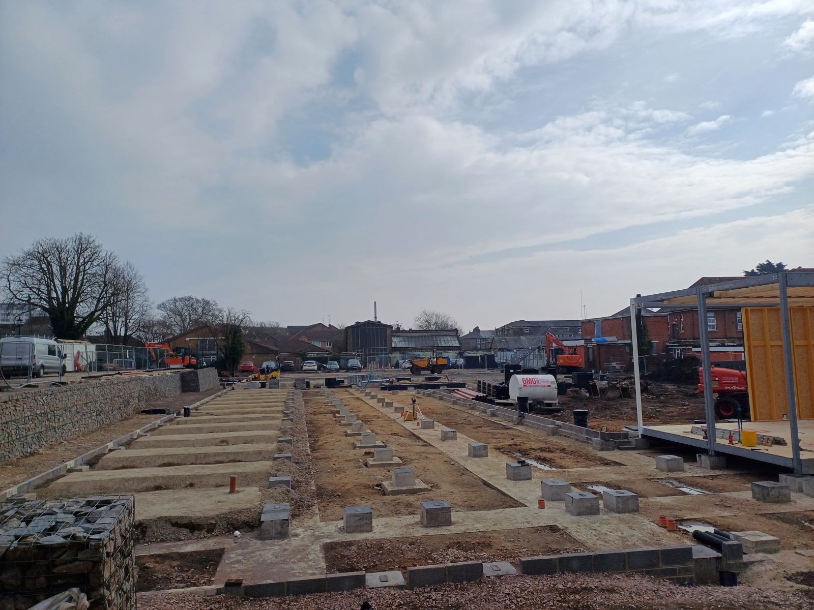 A building site showing a grid of lines of concrete set in the earth as foundations. With a retaining wall to the left made of wire baskets of stones. 