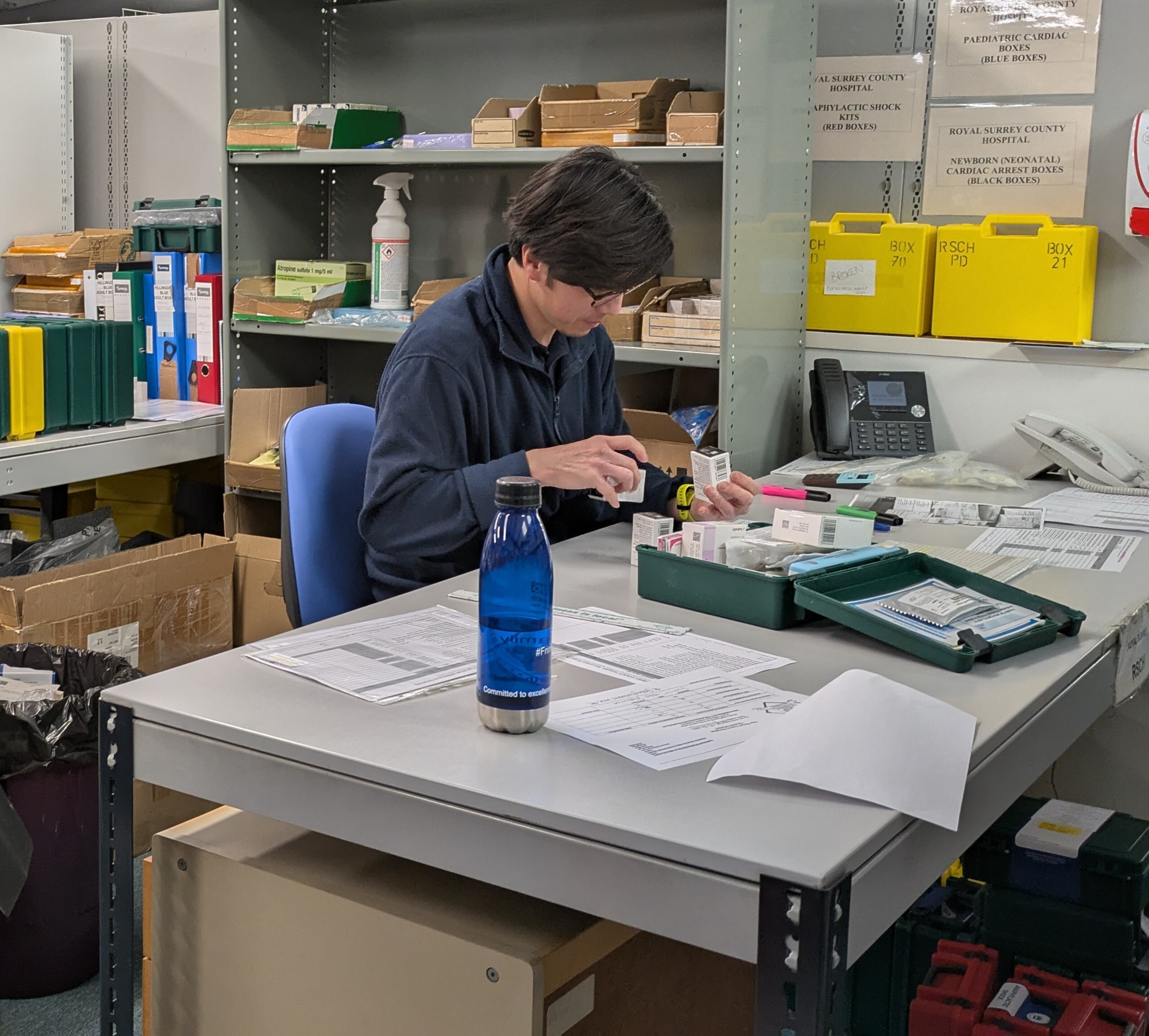 A worker at a cubicle desk in a warehouse type building, sorting drug packets from a first aid kit