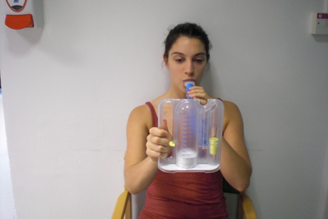 Person seated blowing into spirometer- a plastic device with a handle, a mouth piece, and 2 vertical tubes - one with gradations marked