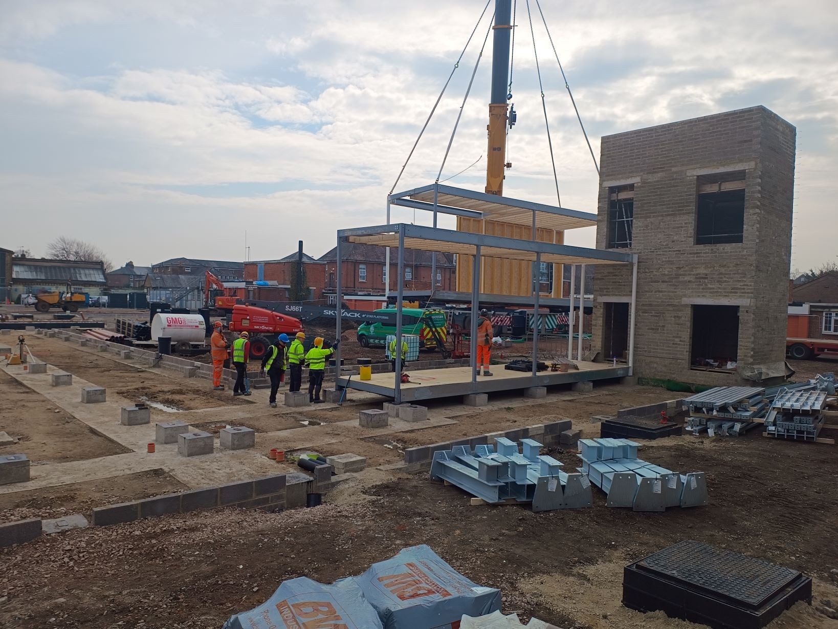 The building site with one module- an open steel frame with a floor- in place on it's foundations, and another being lifted into place by crane